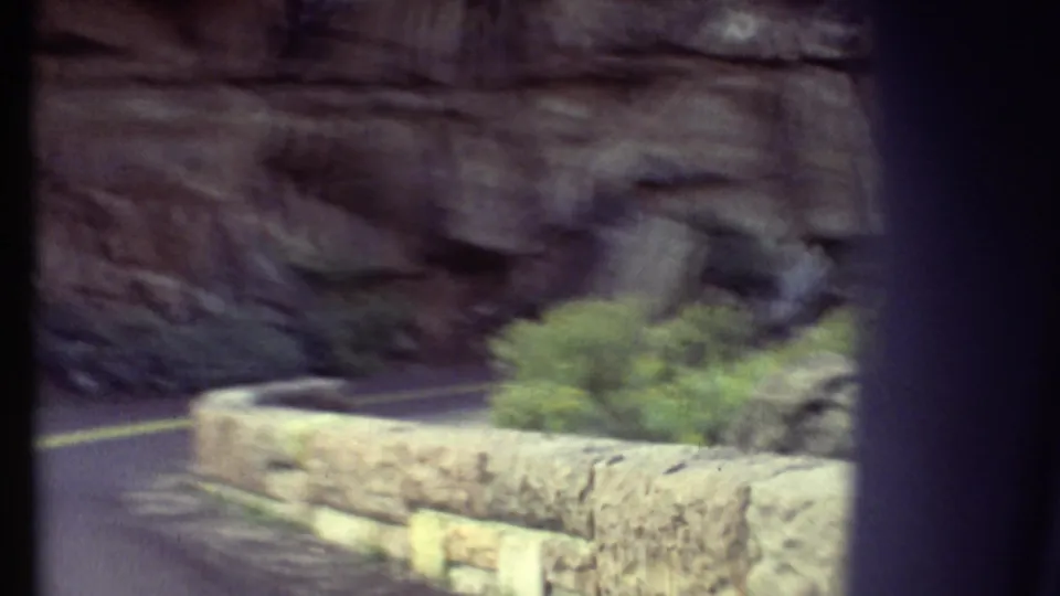 Grand Canyon road footage from 1972 with the camera moving past a stone wall, shrubs, and a cliff-lined curve.