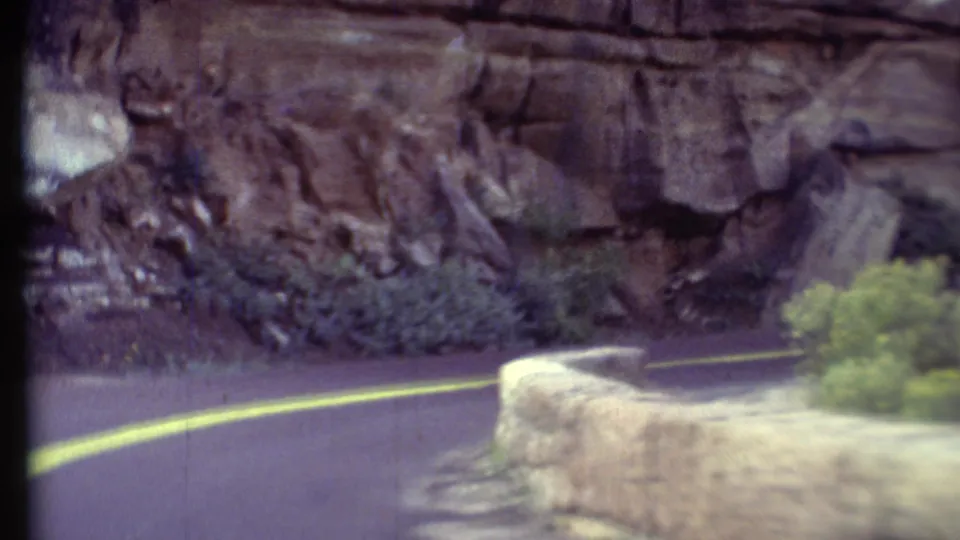 Vintage 8mm view of a Grand Canyon road curve with double yellow markings, a stone barrier, and layered rock nearby.