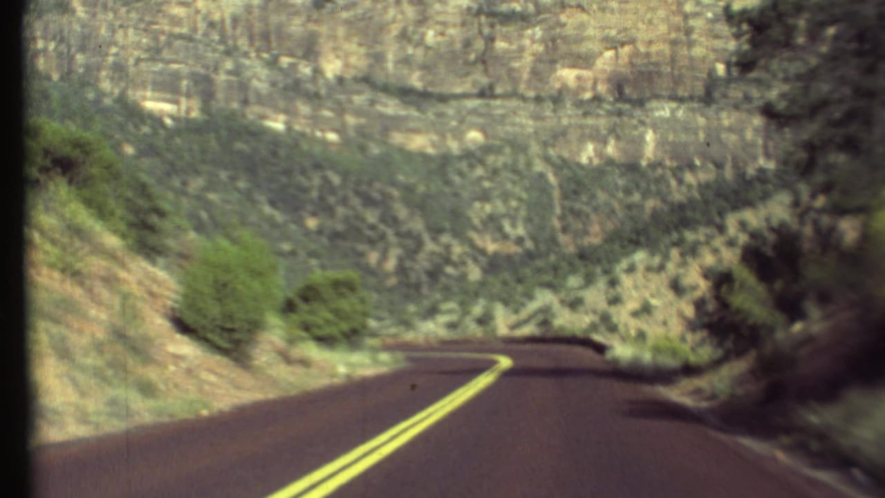 1972 8mm Grand Canyon road view showing a curving two-lane asphalt road, canyon walls, and sparse desert vegetation.
