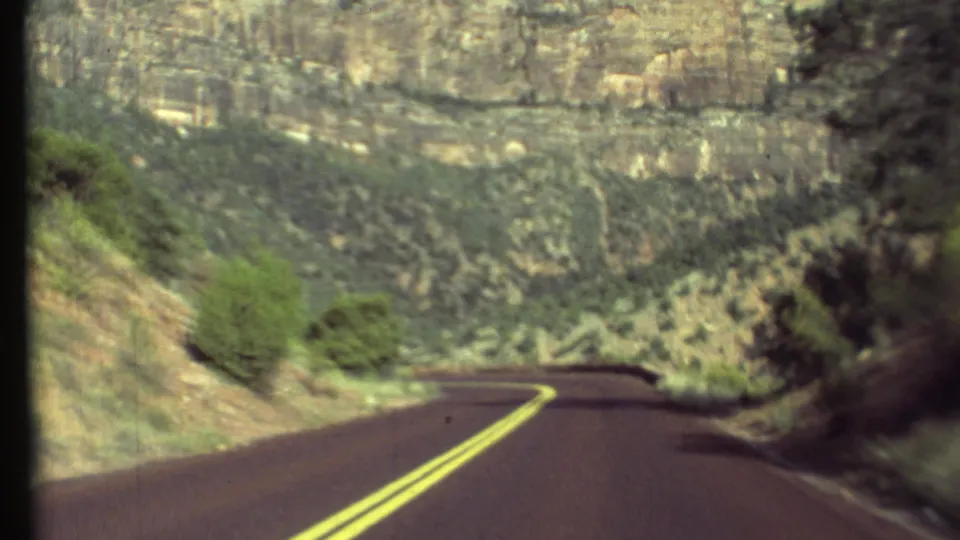 1972 8mm Grand Canyon road view showing a curving two-lane asphalt road, canyon walls, and sparse desert vegetation.