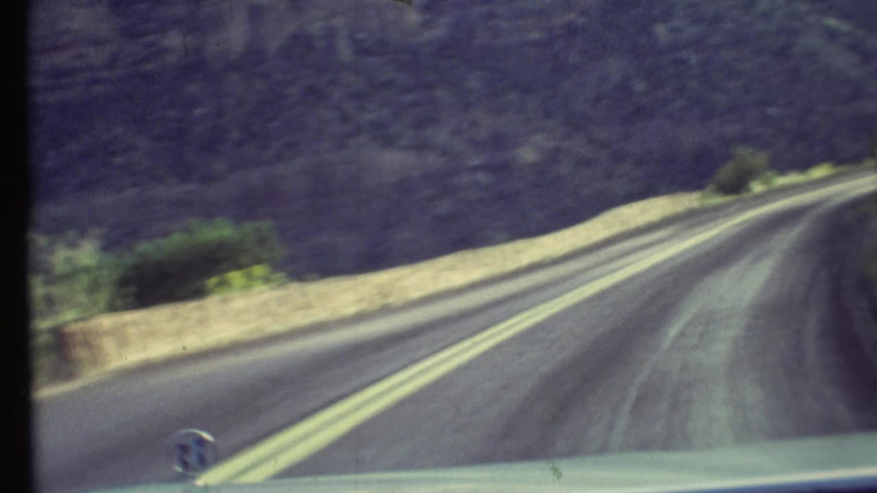 1972 Grand Canyon driving view from inside a car, with a paved road, double yellow lines, low barrier, and canyon slope ahead.