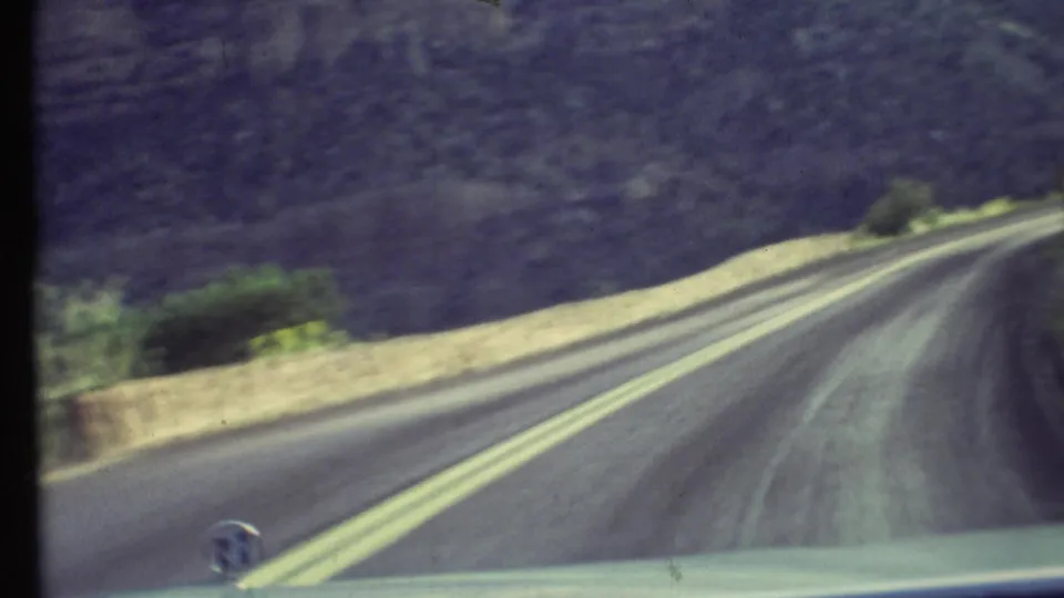 1972 Grand Canyon driving view from inside a car, with a paved road, double yellow lines, low barrier, and canyon slope ahead.