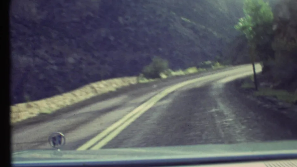 1972 8mm windshield view in Grand Canyon, with the car hood visible as the road curves past a low barrier and vegetation.