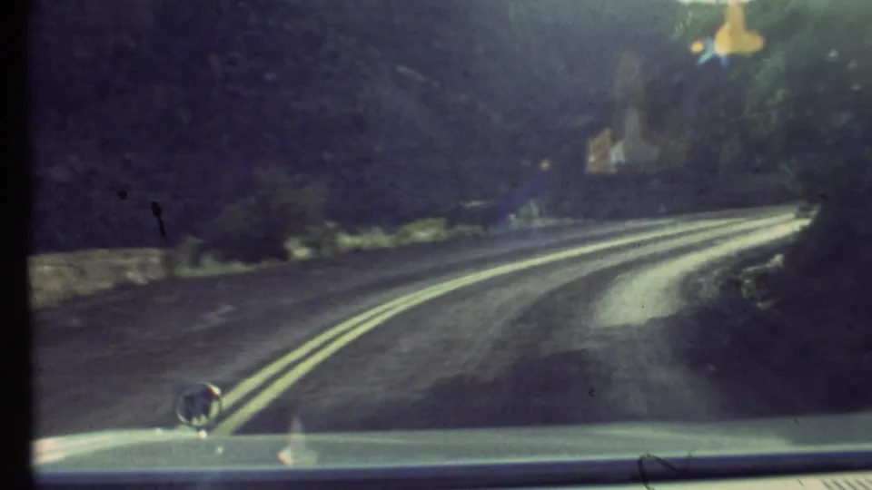 1972 Grand Canyon home-movie frame showing a forward car view along a two-lane road beside canyon walls and sparse vegetation.