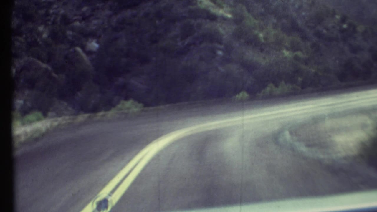 1972 Grand Canyon area dashboard view of a winding road with yellow center lines, roadside vegetation, and soft 8mm blur.