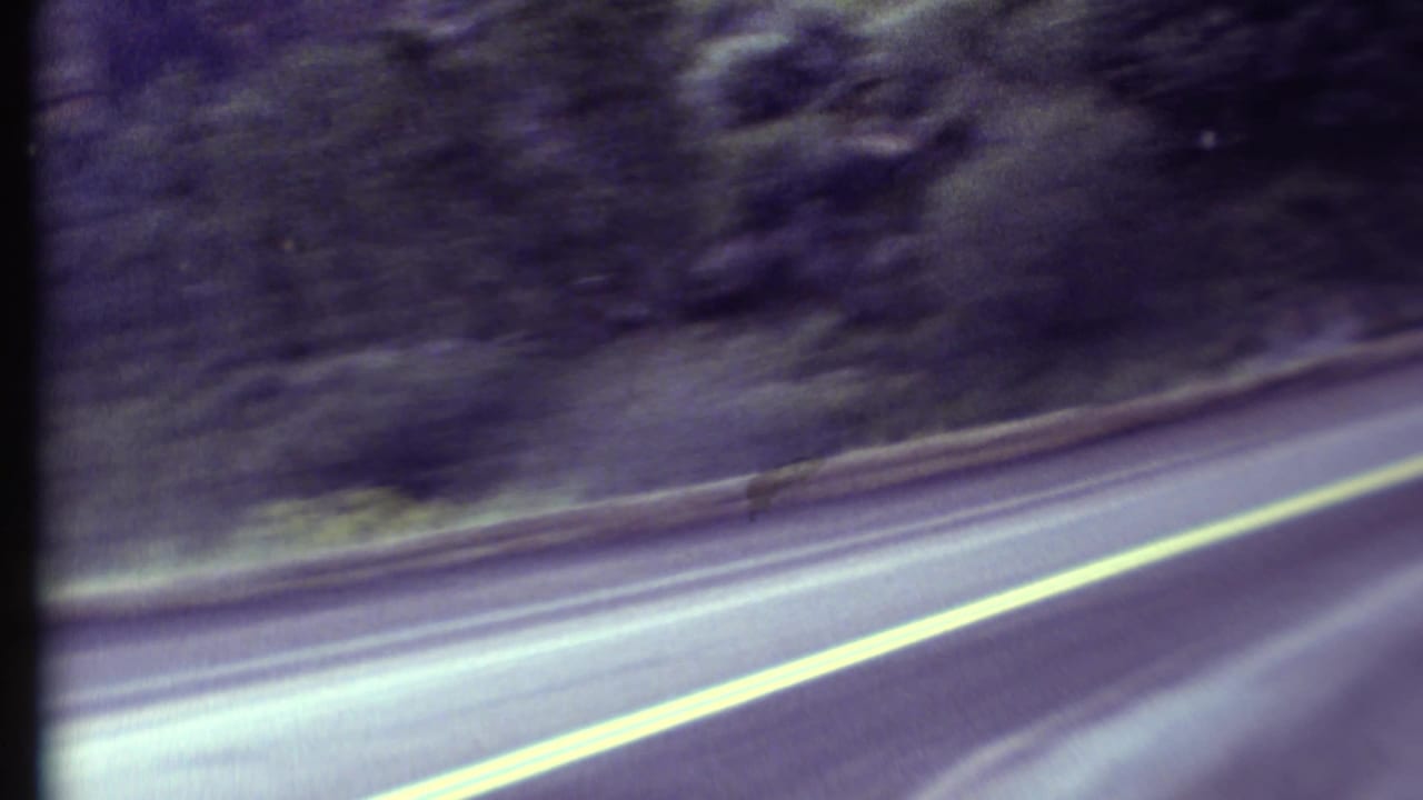 1972 8mm Grand Canyon region road-trip view from a moving car, showing a curving road, yellow center line, and wooded hillside.