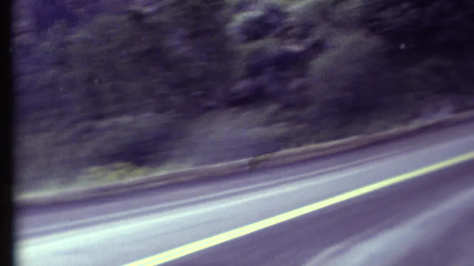 1972 8mm Grand Canyon region road-trip view from a moving car, showing a curving road, yellow center line, and wooded hillside.
