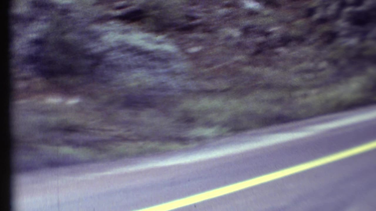 1972 8mm roadside view in the Grand Canyon region, with a yellow center line and trees blurring along a rocky slope.