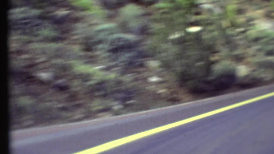 Vehicle-perspective 8mm frame from the 1972 Grand Canyon region trip, showing road edge, yellow line, trees, and rocky hillside.
