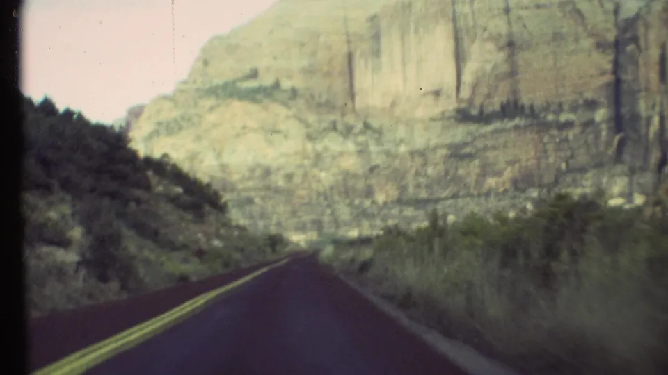 1972 8mm frame looking down a road toward a tall Grand Canyon rock wall, with sparse vegetation along both sides.