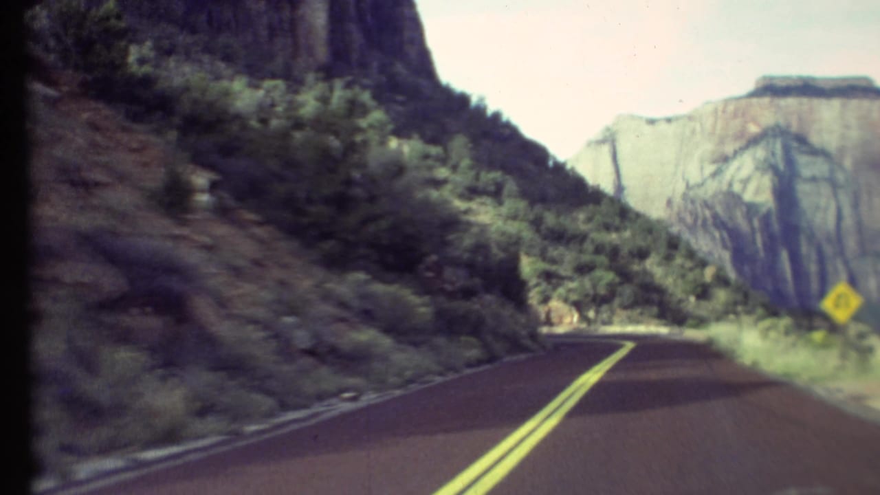1972 8mm Grand Canyon road view with a two-lane bend, steep rock wall, roadside plants, and a pale canyon face ahead.