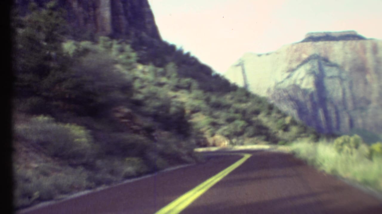 Forward-facing 8mm view of a Grand Canyon road bending between dark roadside rock and a broad pale canyon wall.