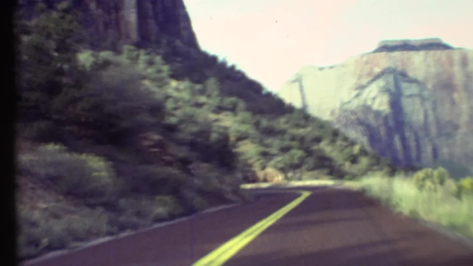 Forward-facing 8mm view of a Grand Canyon road bending between dark roadside rock and a broad pale canyon wall.