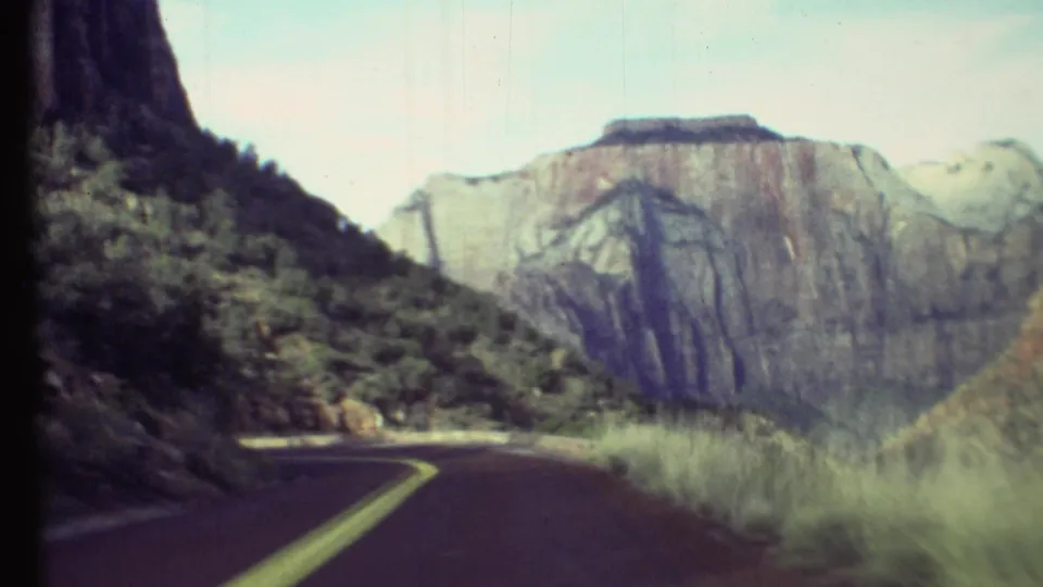 1972 Grand Canyon driving frame showing a bend in the road, roadside grass, dark cliff at left, and large canyon wall beyond.