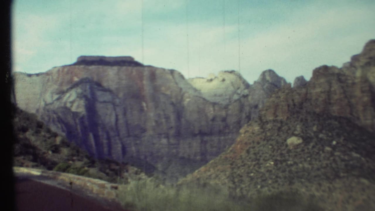 1972 8mm view of Grand Canyon cliffs and desert slopes, framed by dark film gate edges and a pale blue sky.