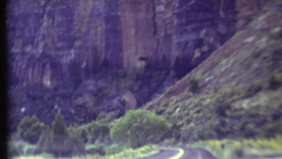 1972 Grand Canyon road view from a moving vehicle, showing a curved paved road, trees, shrubs, and high canyon walls.