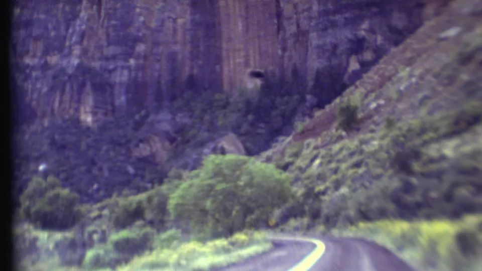 Vintage 8mm Grand Canyon road scene with a curve in the pavement, green shrubs, and towering reddish rock formations.