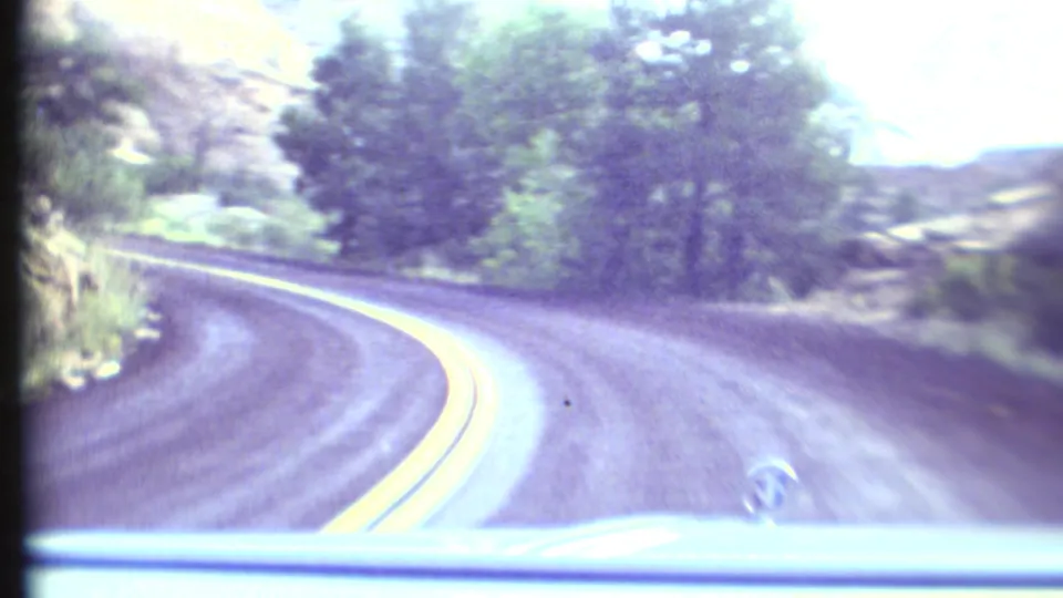 Overexposed 1972 Grand Canyon-area windshield view showing a paved curve, double yellow lines, roadside trees, and rocky terrain.