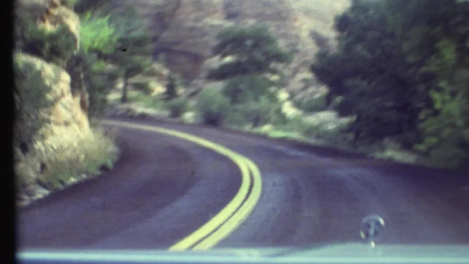 1972 Grand Canyon-area driving view from inside a car, with a winding two-lane road, yellow centerlines, trees, rocks, and a dashboard emblem.