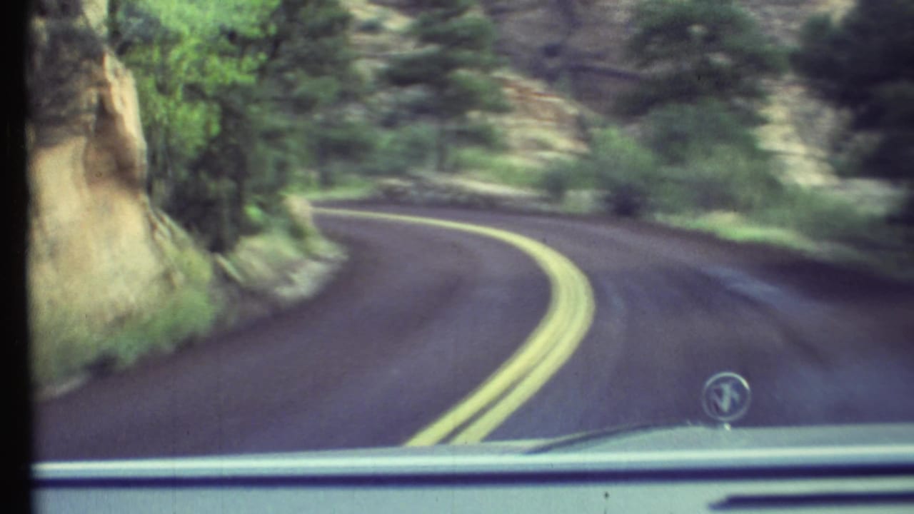 Curving paved road seen through a car windshield, with double yellow lines, trees, rocks, and the car hood visible.