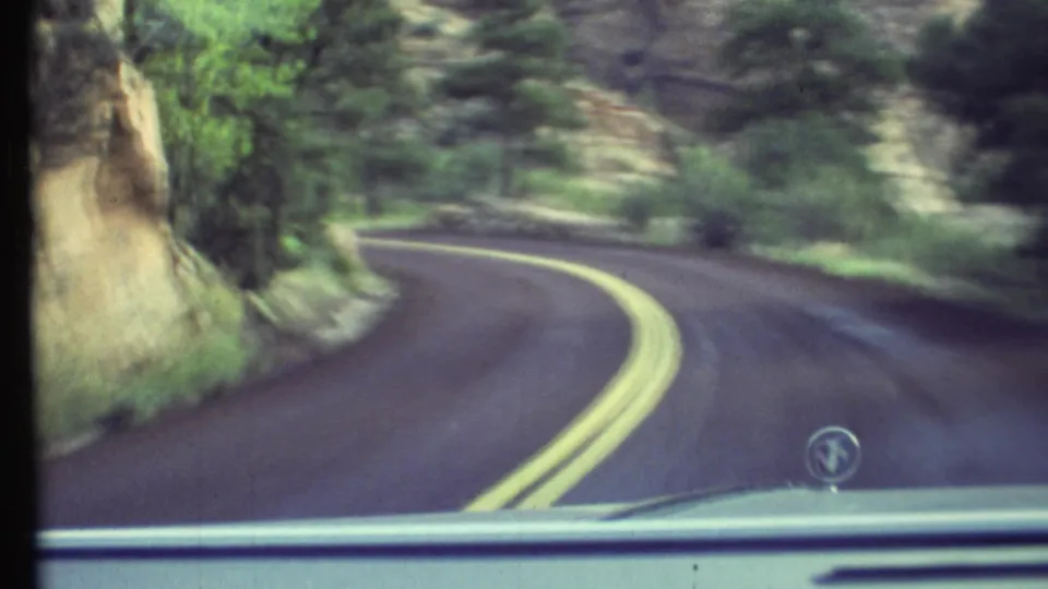 1972 Grand Canyon-area driving view through a windshield, showing a winding forest road, rocky shoulder, double yellow lines, and the car hood.