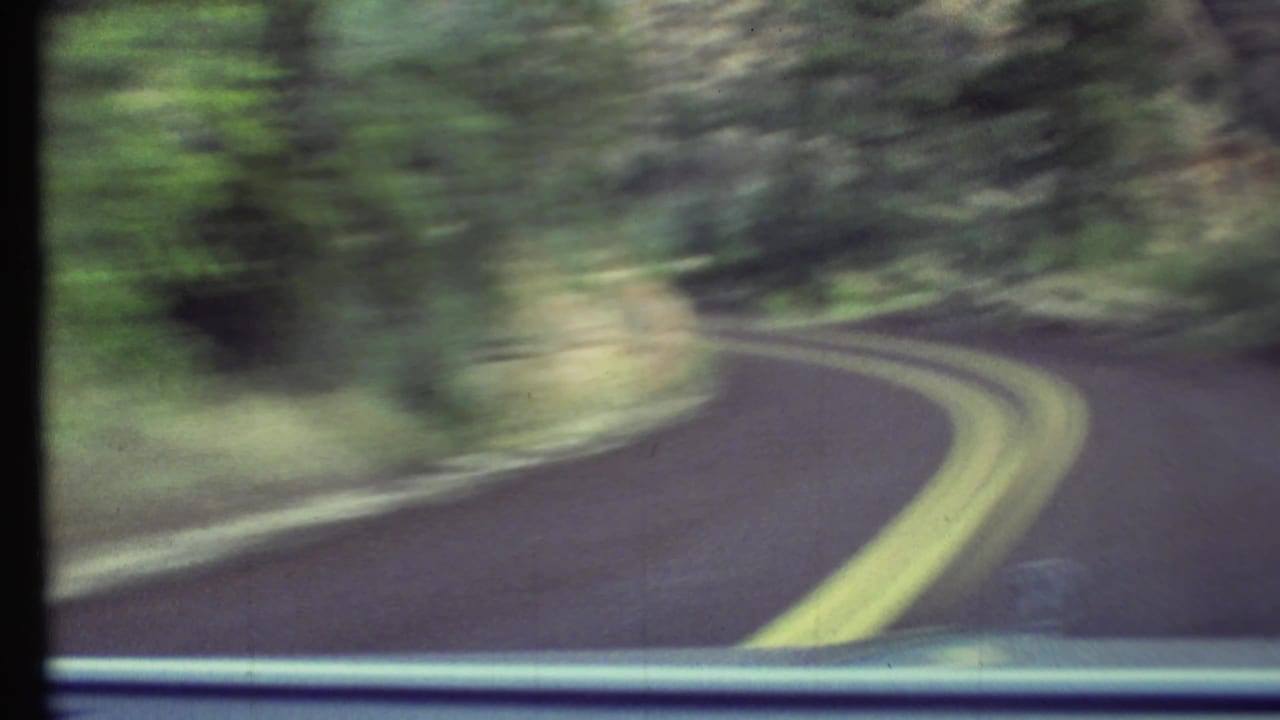 Motion-blurred 1972 in-car view of a winding Grand Canyon-area road bordered by rocks, shrubs, and trees.