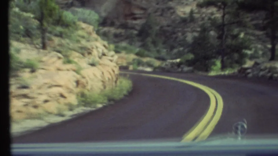 1972 home-movie frame from inside a car, showing a forested canyon road curving past pale rock walls in the Grand Canyon area.