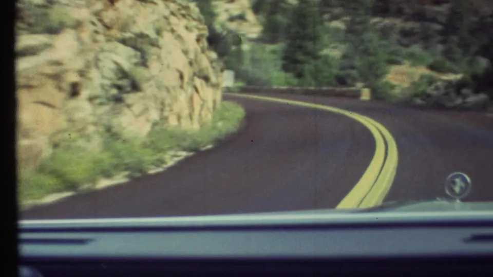 Windshield view from a 1972 Grand Canyon-area drive, looking down a curving road bordered by rock walls and green vegetation.