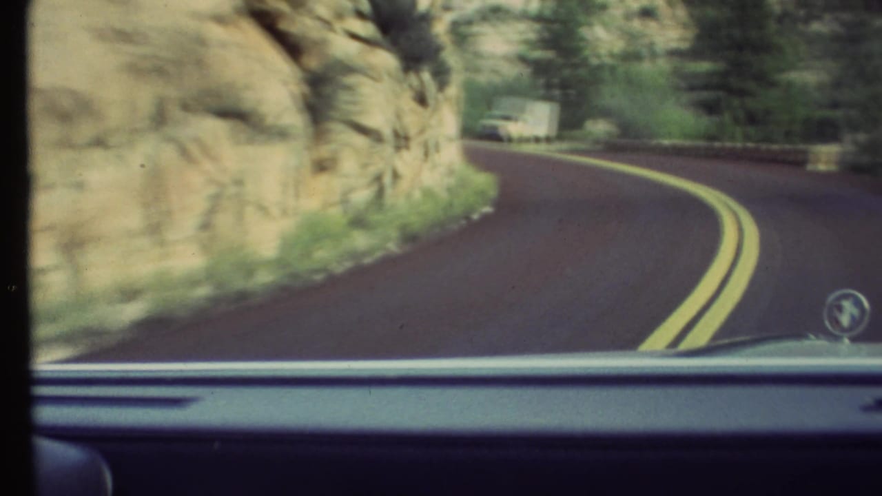 1972 8mm Grand Canyon-area driving view from inside a car, showing a bend in the road, rocky cliff, vegetation, and a white truck ahead.