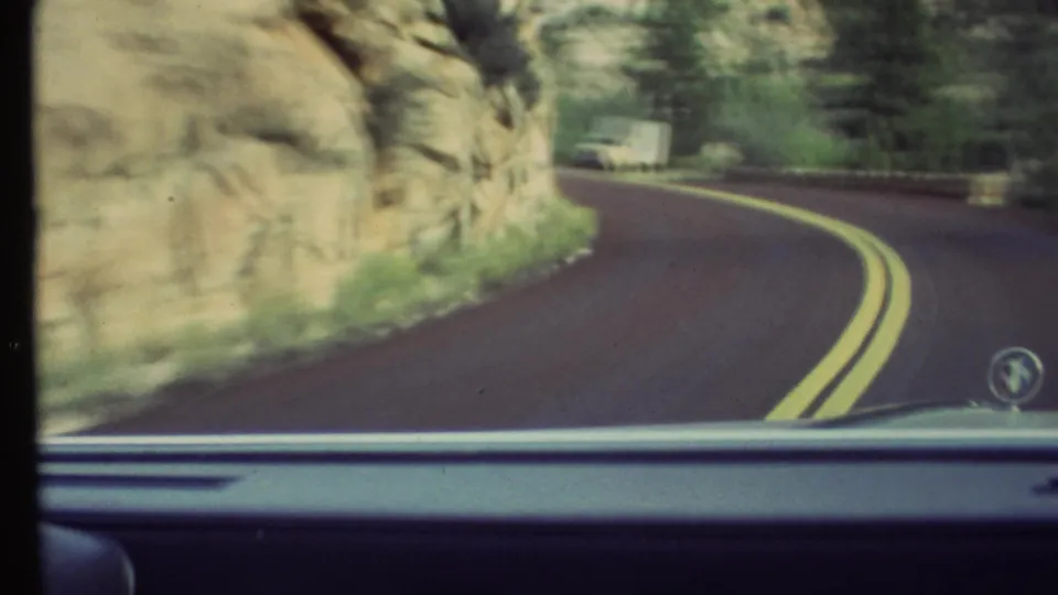 1972 8mm Grand Canyon-area driving view from inside a car, showing a bend in the road, rocky cliff, vegetation, and a white truck ahead.