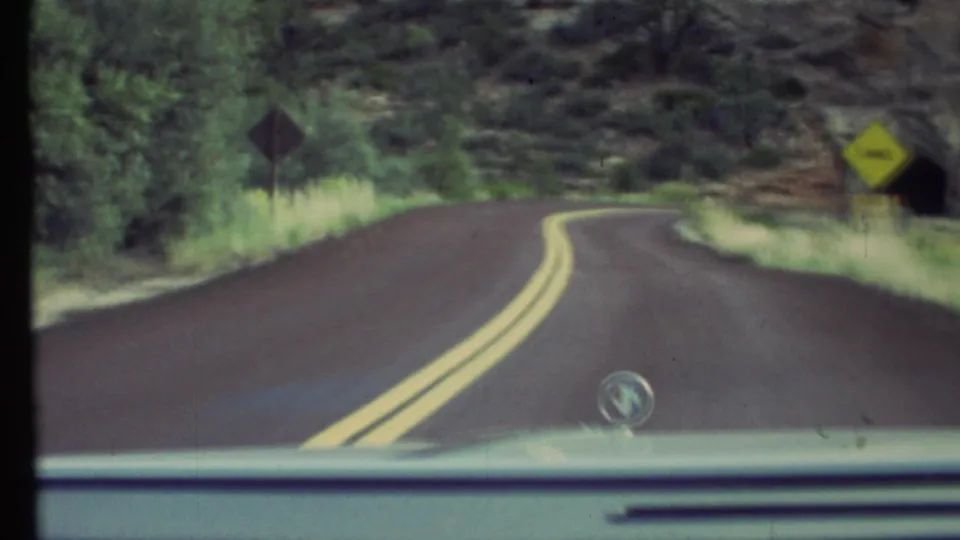 1972 8mm driving view in the Grand Canyon area, showing a curving road, roadside signs, grass, rocky slope, and a dark tunnel opening.