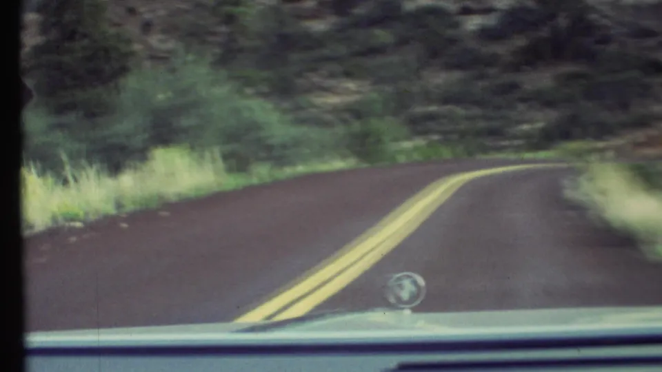 1972 Grand Canyon-area car POV on a winding asphalt road with double yellow lines, roadside grass, green brush, and rocky terrain.