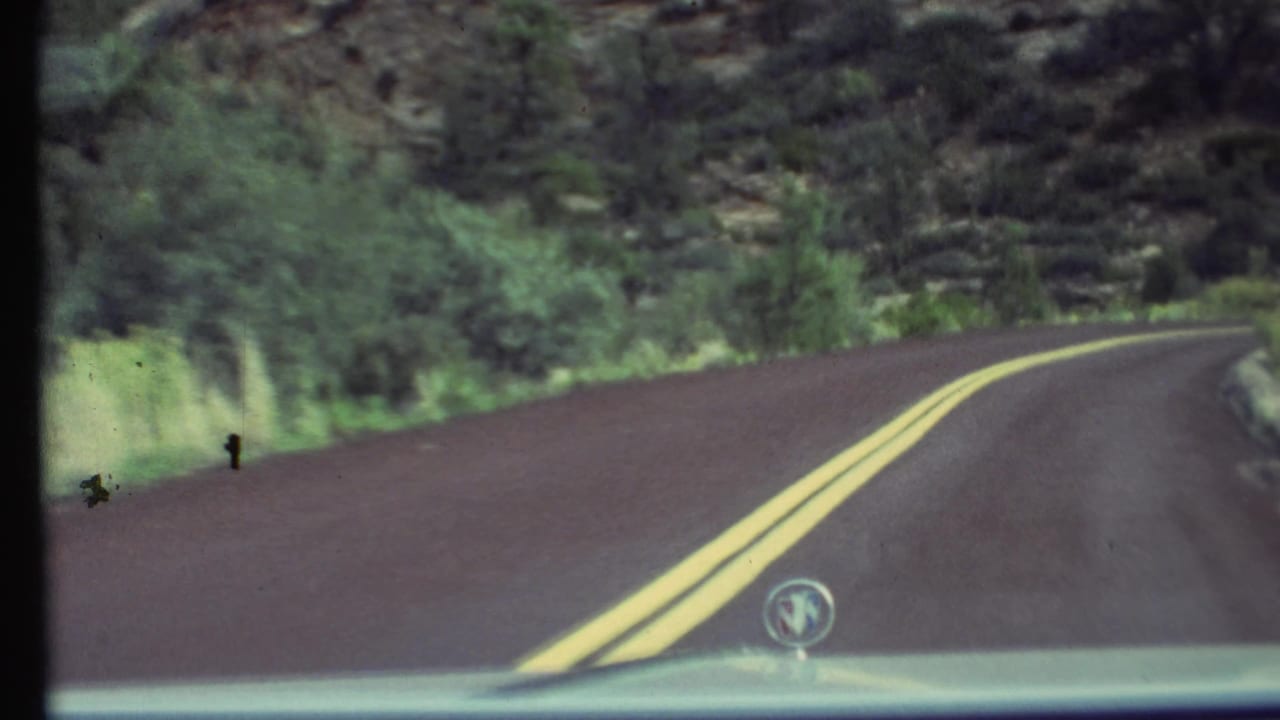 1972 8mm Grand Canyon travel footage showing a winding road, double yellow lines, roadside vegetation, and a hood ornament at the bottom of frame.