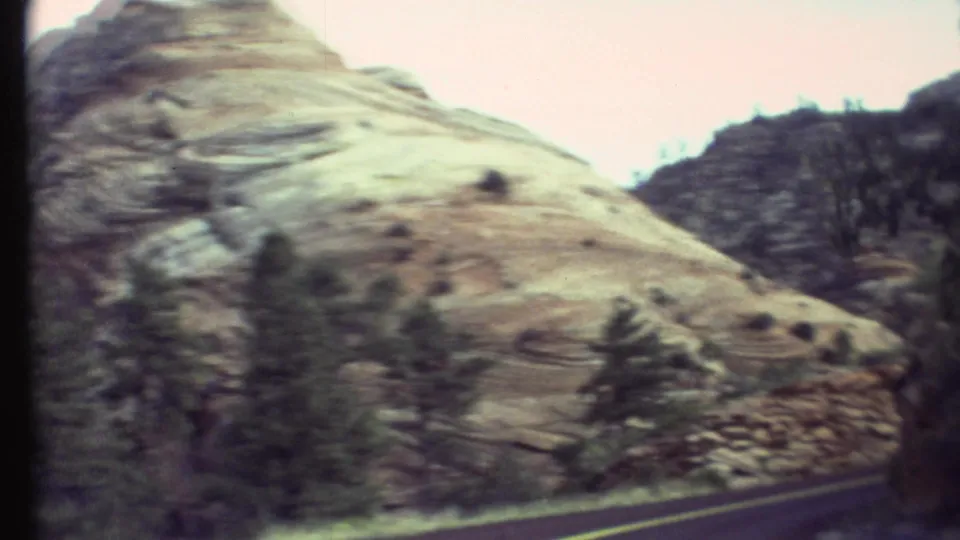 1972 Grand Canyon road-trip frame showing pale layered rock, darker ridge lines, scattered trees, and roadside motion blur from the car.