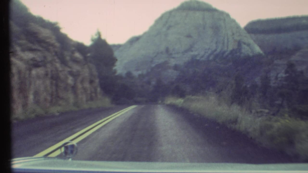 1972 Grand Canyon area driving view with the car hood and side mirror visible below a two-lane road and pale rock formation.