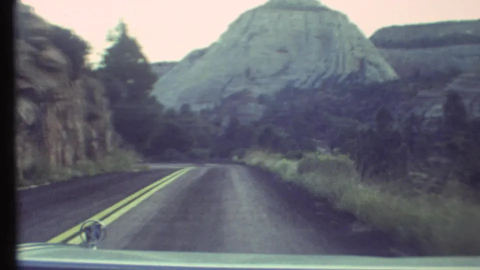 1972 8mm car POV through the Grand Canyon area, showing a two-lane road, side mirror, grasses, and pale canyon rock.