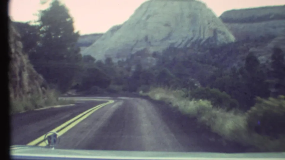 1972 Grand Canyon area road footage, with the car hood framing a curve toward trees and a pale canyon formation.