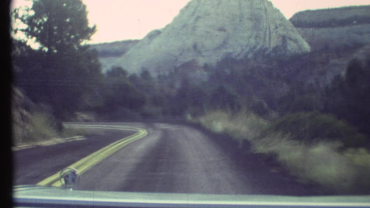 Paved road with double yellow lines curves toward pale rock formations and roadside trees.