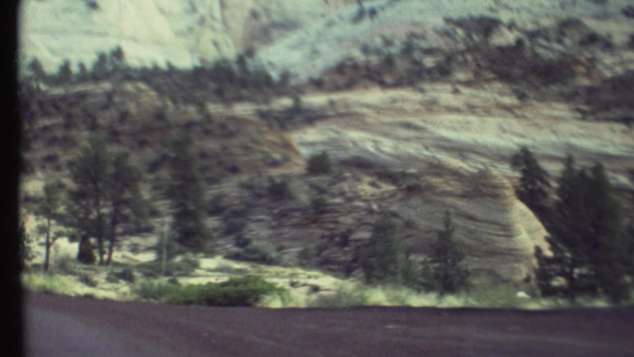 Moving 8mm view of pale layered rock, scattered trees, and roadside terrain in the Grand Canyon region.