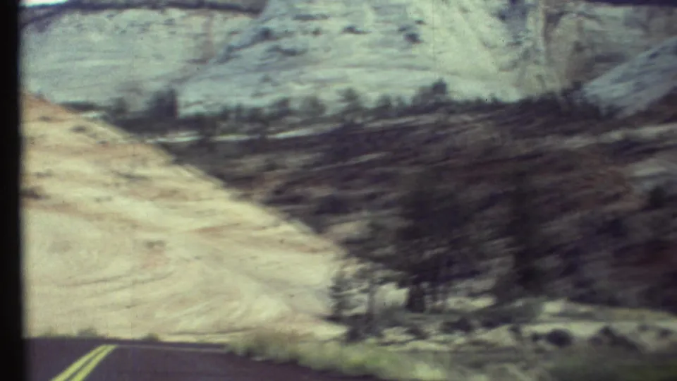 1972 home-movie frame from a moving car, showing road markings, dark trees, and pale eroded slopes in Arizona canyon terrain.