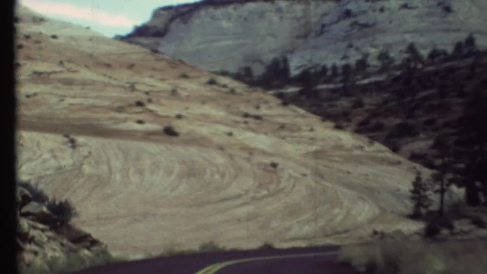 Soft 8mm frame of Grand Canyon region terrain, with a pale sloped hillside, sparse vegetation, trees, and road foreground.