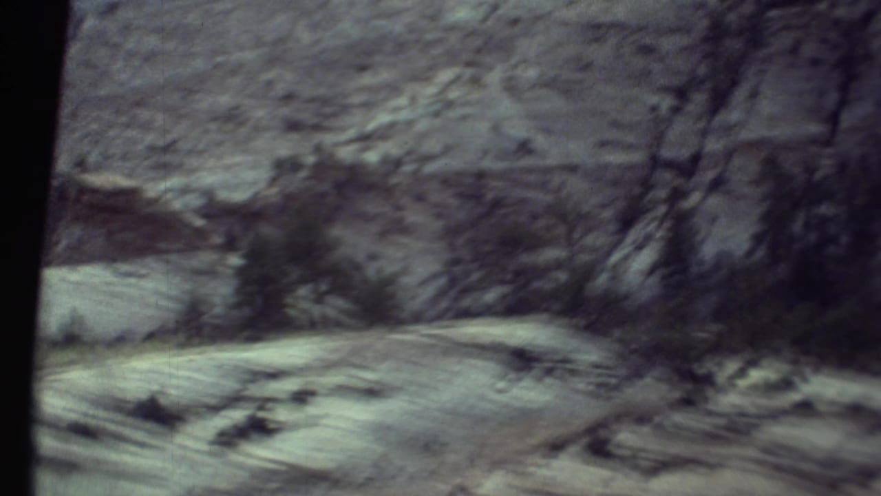 1972 Grand Canyon travel footage showing a blurred rocky wall, sparse trees, and pale roadside ground from inside a moving vehicle.