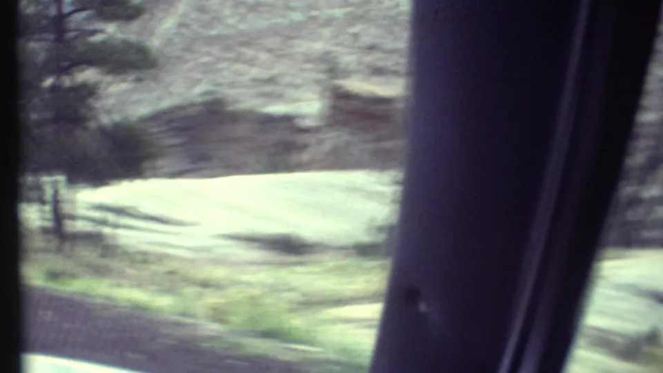 1972 Grand Canyon road footage with pale roadside terrain, rock wall, trees, and a dark vehicle window pillar in the foreground.