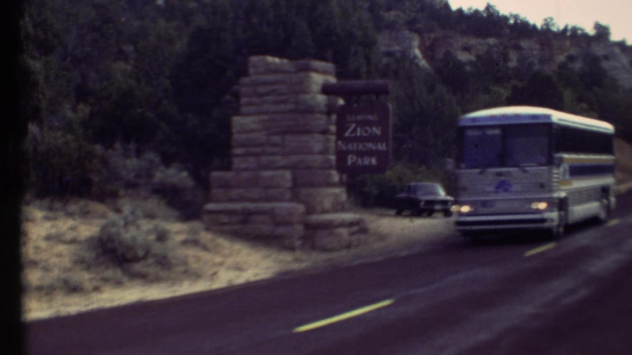 A tour bus drives past the stone Zion National Park entrance sign, with a dark car nearby and rocky wooded hills behind it.