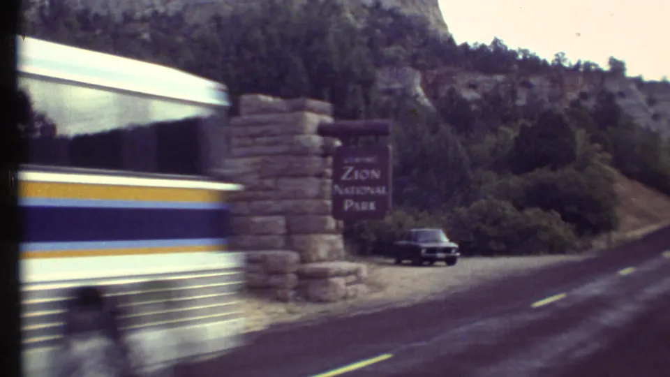 The bus passes close to the camera, creating motion blur as the Zion National Park sign, road, and dark car remain visible in the background.