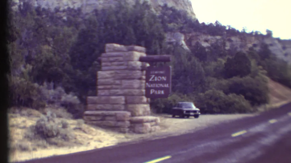 The 1972 8mm view settles on the Zion National Park entrance sign, a dark car, the paved road, roadside brush, and rocky wooded hills.
