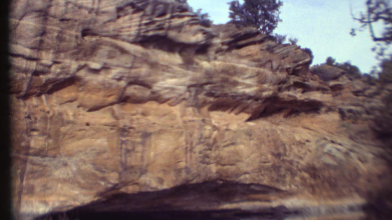 1972 8mm travel frame showing layered desert sandstone cliffs with sparse vegetation and a pale sky in the Grand Canyon region.