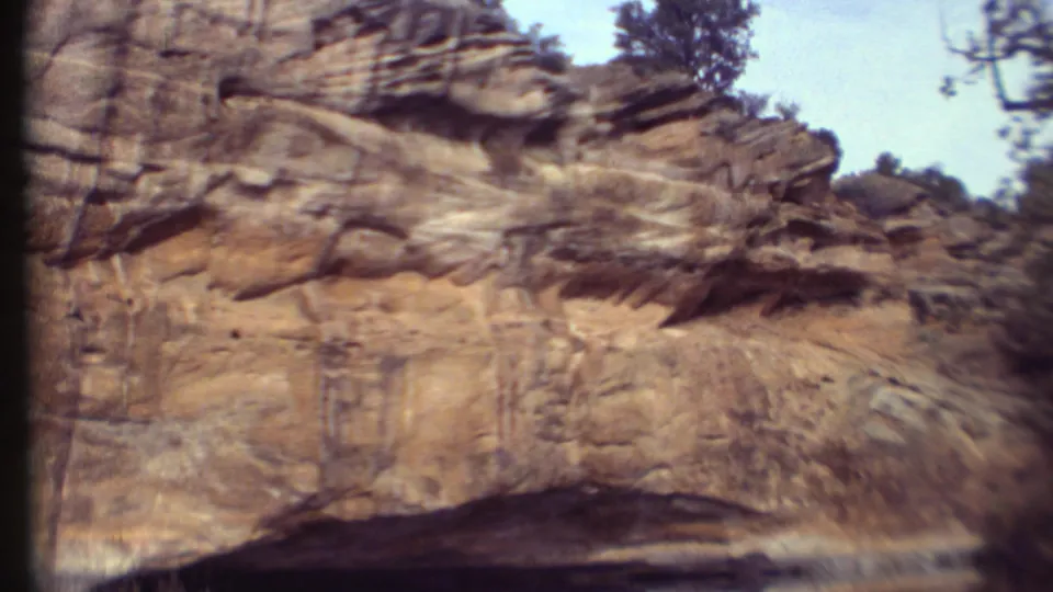 1972 8mm travel frame showing layered desert sandstone cliffs with sparse vegetation and a pale sky in the Grand Canyon region.