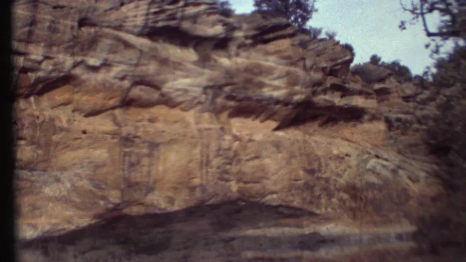 A handheld 8mm frame shows a sandstone cliff face with vertical staining, brush, and shadowed foreground in the Grand Canyon region.