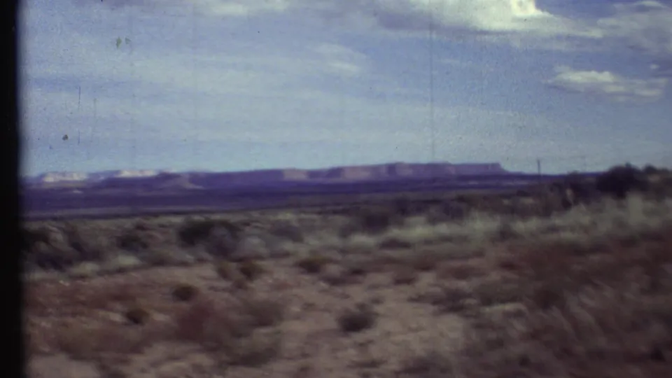 1972 8mm travel footage view across arid scrubland toward distant mesas under a pale, lightly clouded sky.